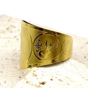 May include: A gold-colored ring with a wide band. The ring features engraved musical symbols, including a treble clef, a guitar, and a heartbeat line. The design is repeated around the band, creating a musical theme.