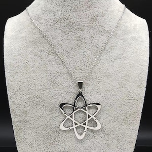 Puede incluir: Un collar de metal en tono plateado con un colgante en forma de estructura atómica con órbitas elípticas que se cruzan. El colgante cuelga de una cadena fina y delicada de color plateado con eslabones pequeños y uniformes, creando un diseño limpio.