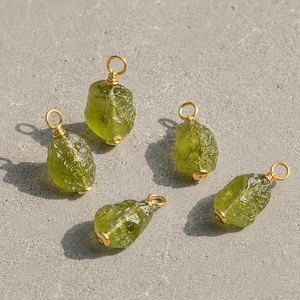 May include: Five raw, translucent olive-green gemstone pendants, each wrapped in thin gold wire with a small circular loop at the top for attachment. The stones have irregular, textured surfaces and are arranged in a scattered formation.