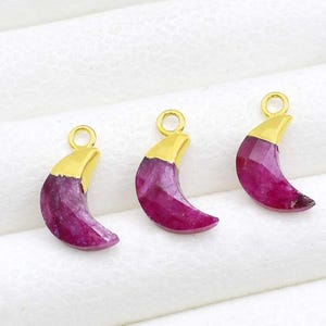 May include: Three ruby crescent moon pendants with gold-coloured caps and loops for jewellery making. The pendants are a deep magenta colour and are displayed on a white surface.