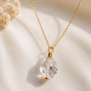 May include: A gold-toned chain necklace featuring a single, irregular, clear, faceted crystal pendant. The crystal is secured at the top and bottom with gold-toned wire wrapping, creating a simple, dangling focal point on a delicate metal chain.