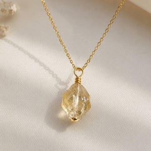 Può includere: Una collana a catena color oro con un ciondolo in pietra preziosa gialla grezza e traslucida. La pietra è fissata con un filo metallico color oro avvolto sulla parte superiore, che la collega alla delicata catena a maglie che pende.