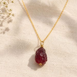 Op de afbeelding: Een gouden ketting met een enkele, ruwe, dieprode granaathanger. De steen heeft een getextureerd, onregelmatig oppervlak en is bevestigd met een klein gouden oogje. De ketting wordt getoond tegen een lichte, getextureerde stoffen achtergrond.