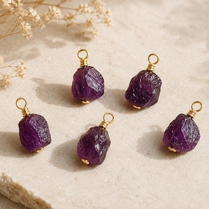 May include: Five raw, deep purple amethyst stone pendants, each featuring a gold-toned wire-wrapped bail at the top. The stones have irregular, faceted surfaces and are arranged in a loose cluster, showcasing their natural, unpolished texture.