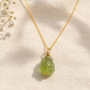 Può includere: Una collana a catena color oro con un ciondolo in pietra grezza verde chiaro. La pietra presenta una superficie irregolare e ruvida, fissata alla sottile catena metallica tramite un piccolo avvolgimento in filo dorato e un anellino.