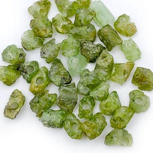 May include: A collection of raw, uncut peridot gemstones in various shapes and sizes. The stones display a range of green hues, from light to olive, with a natural, rough texture. The gemstones are scattered on a white surface.