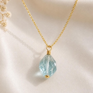 May include: A gold-toned chain necklace featuring a single, faceted, light blue gemstone pendant. The stone is secured with gold-toned wire wrapping at the top and a small gold-toned bead at the bottom, creating a delicate and refined jewellery piece.