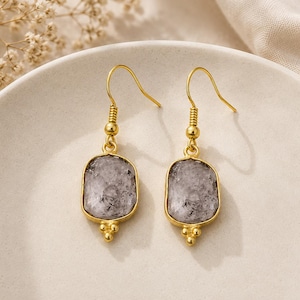 May include: A pair of gold-toned drop earrings featuring rectangular, faceted, translucent gray stones set in metal bezels. Each earring has a gold hook wire and a small decorative cluster of three tiny gold spheres at the bottom edge.