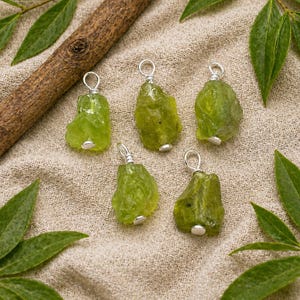 Può includere: Cinque ciondoli di pietre preziose di peridoto grezzo con anelli in filo d'argento. Le pietre verde oliva sono esposte su una superficie di lino beige, con un bastoncino di legno e foglie verdi. I ciondoli sono pronti per essere indossati come collane.