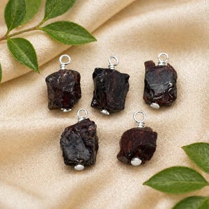 Può includere: Cinque ciondoli di granato rosso scuro grezzo con anelli color argento. Le pietre preziose grezze sono esposte su un tessuto beige, con foglie verdi sullo sfondo. Ogni ciondolo misura circa 2 cm di lunghezza.