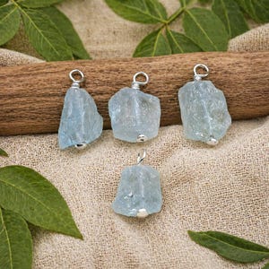 Può includere: Quattro ciondoli in acquamarina con anelli argentati. Le pietre preziose grezze, di colore azzurro chiaro, sono esposte su un tessuto beige con un tronco di legno e foglie verdi sullo sfondo. Ogni ciondolo ha una piccola perla d'argento sul fondo.