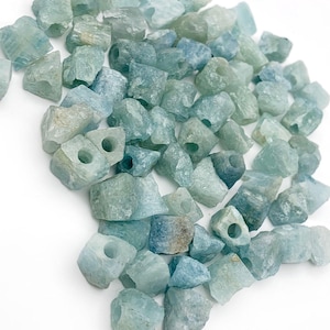 Op de afbeelding: Een verzameling ruwe aquamarijn edelsteen kralen. De kralen zijn onregelmatig van vorm en hebben een ruwe textuur. De kleur varieert van lichtblauw tot dieper blauwgroen, met enkele bruine tinten. Elke kraal heeft een gat.