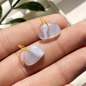 May include: Two cloud-shaped earrings. The earrings are made of a translucent, pale lavender material with white marbling. Each earring has a small, gold-colored hoop at the top. The earrings are displayed on a person's hand.