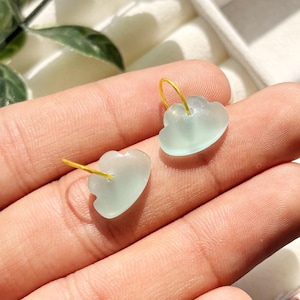 May include: A pair of cloud-shaped earrings. Each earring features a pale green, translucent cloud charm suspended from a gold-colored hoop. The earrings are displayed against a neutral background, highlighting their delicate design and color.