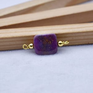 May include: A square, purple gemstone bead with gold-colored accents. The bead has a textured surface and is connected to gold-colored loops on either side. The background includes wooden elements and a white surface.