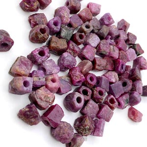 May include: A collection of raw, uncut ruby beads in various shades of red and pink. Each bead has a hole drilled through the center, ready for stringing. The gemstones have a rough, natural texture and are scattered on a white surface.