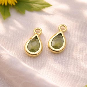 Può includere: Due ciondoli a goccia color oro con una pietra verde al centro. I ciondoli hanno un piccolo anello nella parte superiore per appenderli. Sfondo bianco con un fiore giallo e foglie verdi.