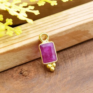 May include: A gold-colored pendant with a rectangular, magenta-colored gemstone. The pendant has a small loop at the top and three small, round gold accents at the bottom. The pendant is set against a wooden surface.
