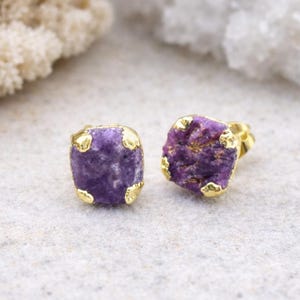 Op de afbeelding: Een paar vierkante oorstekers. Elke oorbel heeft een ruw geslepen, dieppaarse steen, gezet in een goudkleurig metalen frame. De oorbellen worden gepresenteerd op een lichtgrijze ondergrond.