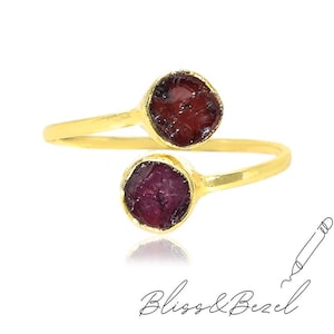 May include: A gold-colored ring with two raw, dark red gemstones. The ring band curves around the finger, with each stone set in a bezel. The text "Bliss & Bezel" is written in cursive below the ring.