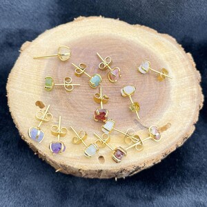 May include: A collection of gold-colored stud earrings with various colorful gemstones, including green, pink, and purple. The earrings are displayed on a round, light brown wooden surface.