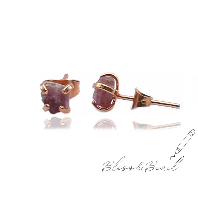 May include: Rose gold stud earrings featuring raw, uncut pink gemstones. The earrings have a simple, elegant design, with the gemstones held in place by a delicate setting. The earrings are displayed on a white background. The brand name "Bliss & Bezel" is written in cursive.