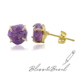 Peut inclure: Boucles d'oreilles à clous avec des pierres précieuses violettes brutes serties dans du métal doré. Les boucles d'oreilles sont présentées sur un fond blanc avec le texte "Bliss & Bezel" en écriture cursive.
