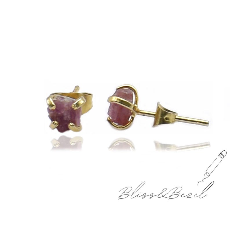 May include: A pair of gold-colored stud earrings, each featuring a raw, pink gemstone. The gemstones have a rough, natural texture. The earrings are displayed on a white background, with the brand name "Bliss & Bezel" in cursive script.
