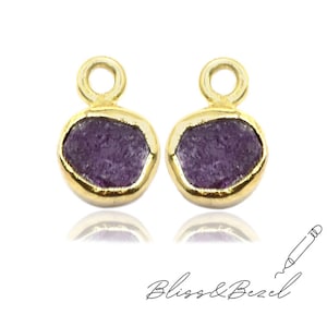 May include: Two gold-colored bezel set charms with a round, rough-cut, purple stone. Each charm has a small loop at the top. The charms are displayed on a white background. The text "Bliss & Bezel" is written in cursive.