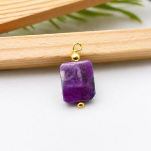 Può includere: Un ciondolo rettangolare in pietra preziosa viola con dettagli dorati. Il ciondolo è di un colore viola intenso con inclusioni più scure. I dettagli dorati includono un piccolo anello nella parte superiore e una perla nella parte inferiore.