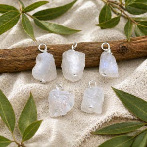 Può includere: Cinque ciondoli grezzi di pietra di luna, azzurri, con anelli in filo d'argento e piccole perline d'argento. I ciondoli sono esposti su un tessuto beige chiaro con foglie verdi e un bastoncino di legno marrone.