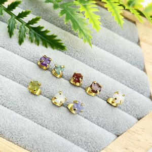 May include: A collection of gold-colored stud earrings, each featuring a different rough-cut gemstone. The gemstones vary in color, including purple, green, red, and white. The earrings are displayed on a gray velvet surface.