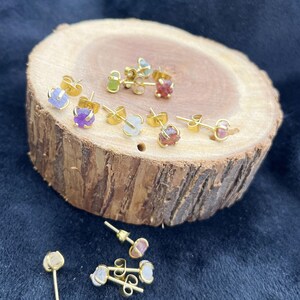 May include: A collection of gold-colored stud earrings with various colorful gemstones, including purple, green, and red. The earrings are displayed on a rustic wooden surface, showcasing their natural beauty and unique designs. The earrings are small and delicate.