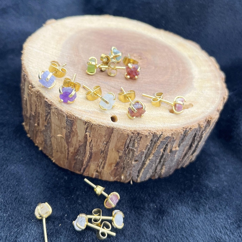 May include: A collection of gold-colored stud earrings with various colorful gemstones, including purple, green, and red. The earrings are displayed on a rustic wooden surface, showcasing their natural beauty and unique designs. The earrings are small and delicate.