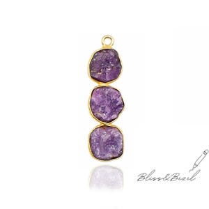 Può includere: Un pendente verticale con tre pietre di ametista viola grezze incastonate in castoni color oro. Le pietre hanno una forma irregolare e una superficie strutturata. Il pendente ha un piccolo anello nella parte superiore per appenderlo. Le parole "Bliss & Bezel" sono scritte in corsivo.
