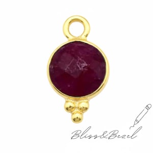 Puede incluir: Un colgante dorado con una gema roja oscura, redonda y facetada. El colgante tiene un lazo en la parte superior y tres pequeñas cuentas doradas debajo. El texto "Bliss & Bezel" está escrito en cursiva.