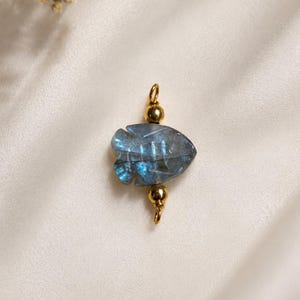 Op de afbeelding: Een sieraadhanger met een gesneden grijze labradorietsteen in de vorm van een vis met iriserende blauwe flitsen. De steen wordt geflankeerd door twee gepolijste goudkleurige metalen kralen en heeft een kleine gouden ring aan de boven- en onderkant.