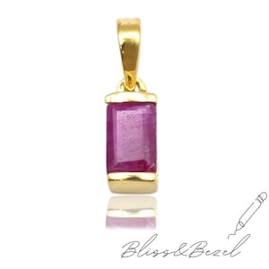 May include: A gold pendant with a rectangular, magenta-colored gemstone. The pendant has a simple, elegant design with a polished finish. The gemstone is set in a gold bezel. The words "Bliss & Bezel" are written in cursive at the bottom.
