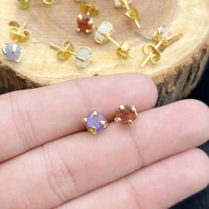May include: Gold-colored stud earrings with raw, uncut gemstones. One pair features a purple stone, the other a reddish-brown stone. Additional earrings with various colored stones are visible in the background.