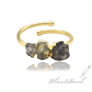 Può includere: Un anello regolabile color oro con tre pietre grezze grigie e nere. L'anello ha un design minimalista con una fascia curva e estremità aperte. Le pietre sono incastonate in un metallo color oro.