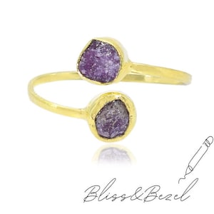 Puede incluir: Un anillo dorado con dos piedras preciosas moradas en bruto. El anillo tiene un diseño abierto, con las piedras engastadas en biseles. El anillo está sobre un fondo blanco. El texto "Bliss & Bezel" está en la parte inferior.
