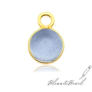 Puede incluir: Un colgante dorado con un centro circular azul claro. El colgante tiene un pequeño lazo en la parte superior para sujetarlo a una cadena o pulsera. Las palabras "Bliss & Bezel" están escritas en cursiva en la parte inferior.