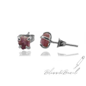 May include: A pair of stud earrings featuring rough-cut, pink gemstones set in silver metal. The earrings have a natural, organic appearance. The image is set against a white background. The brand name "Bliss & Bezel" is written in cursive at the bottom.