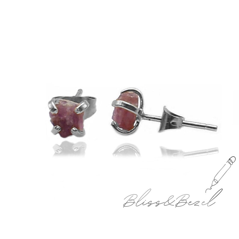 May include: A pair of stud earrings featuring rough-cut, pink gemstones set in silver metal. The earrings have a natural, organic appearance. The image is set against a white background. The brand name "Bliss & Bezel" is written in cursive at the bottom.