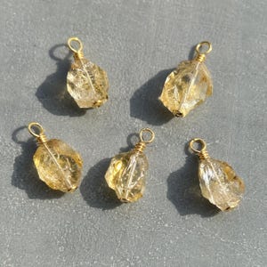 May include: Five raw, translucent yellow citrine crystal pendants, each wrapped in gold-coloured wire with a small circular loop at the top for attachment. The stones have irregular, faceted shapes and are arranged in a scattered formation.