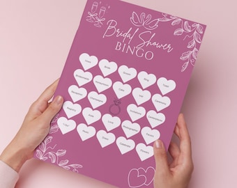 May include: A pink bridal shower bingo game card with white heart-shaped spaces and wedding-themed words. The card features the text "Bridal Shower Bingo" and decorative elements like champagne glasses and a ring.