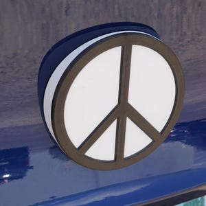 May include: A peace sign emblem with a white center and a dark brown outline. The emblem is layered with a white and dark blue border. The peace symbol is mounted on a blue surface.