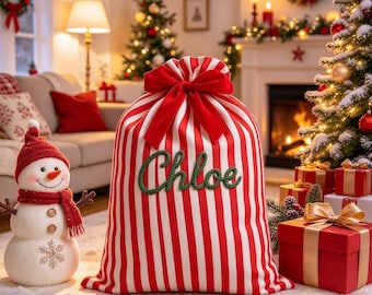 May include: A large fabric gift sack featuring vertical red and white stripes, cinched at the top with a large red velvet bow. The name Chloe is embroidered in dark green script across the center of the sack. The sack sits on a white surface.