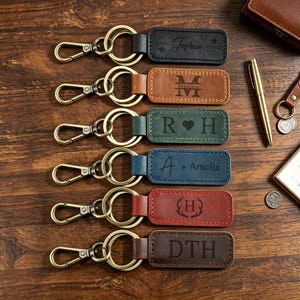 May include: Six rectangular leather keychains with metal lobster clasps and split rings. The keychains are black, tan, green, blue, red, and dark brown, each featuring unique laser-engraved names, initials, or symbols like stars and antlers.