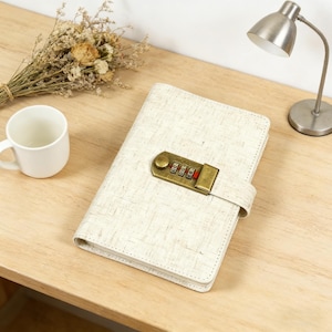 Op de afbeelding: Crèmekleurig notitieboek met een combinatieslot. Het notitieboek heeft een getextureerde kaft en een messingkleurig slot. Een witte mok en een klein boeket droogbloemen zijn ook zichtbaar.